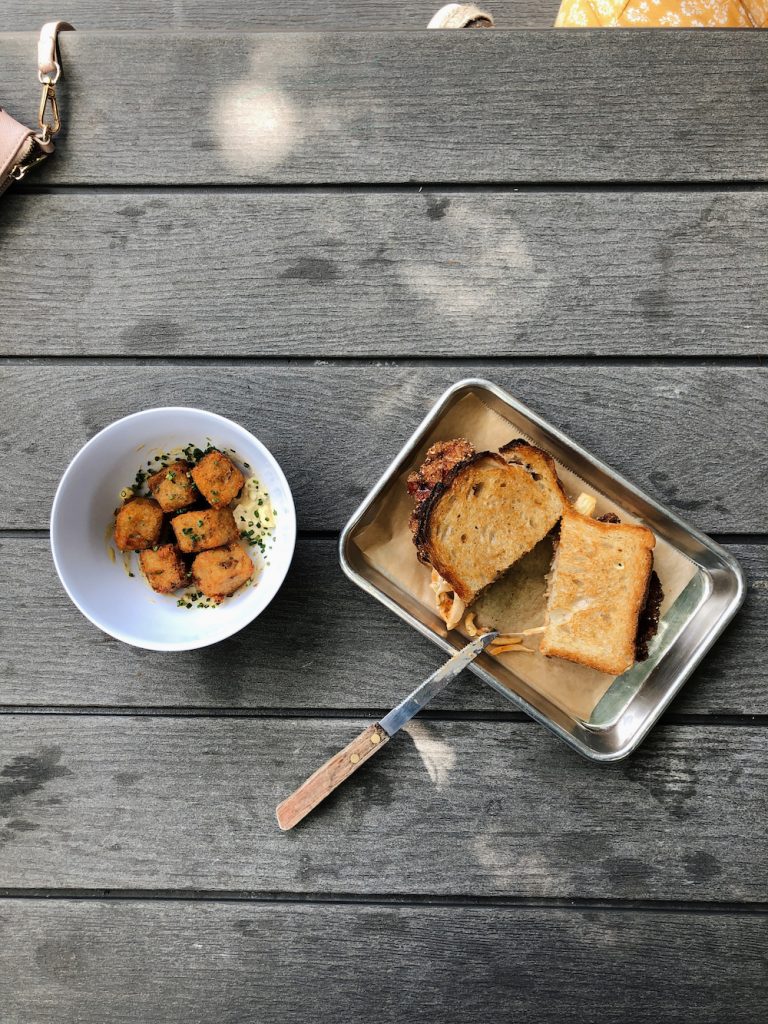 Fried chicken sandwich on metal tray and bowl of tater tots on picnic table at Sour Duck Market in Austin, Texas.