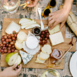 Cheeseboard with pear slices, grapes, honey, and different cheeses on newspaper-covered table.