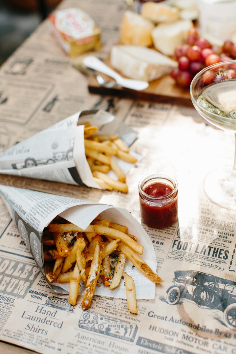 French fries in a newspaper cone on a newspaper-covered table next to a small bottle of ketchup.