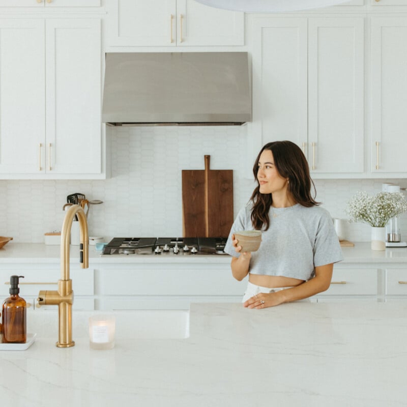 Brunette woman drinking matcha in all-white kitchen.