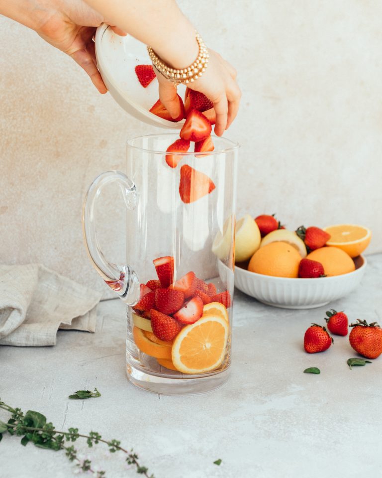 A woman's hand drops strawberries into a pitcher for a rosé sangria recipe next to a bowl of oranges and strawberries.