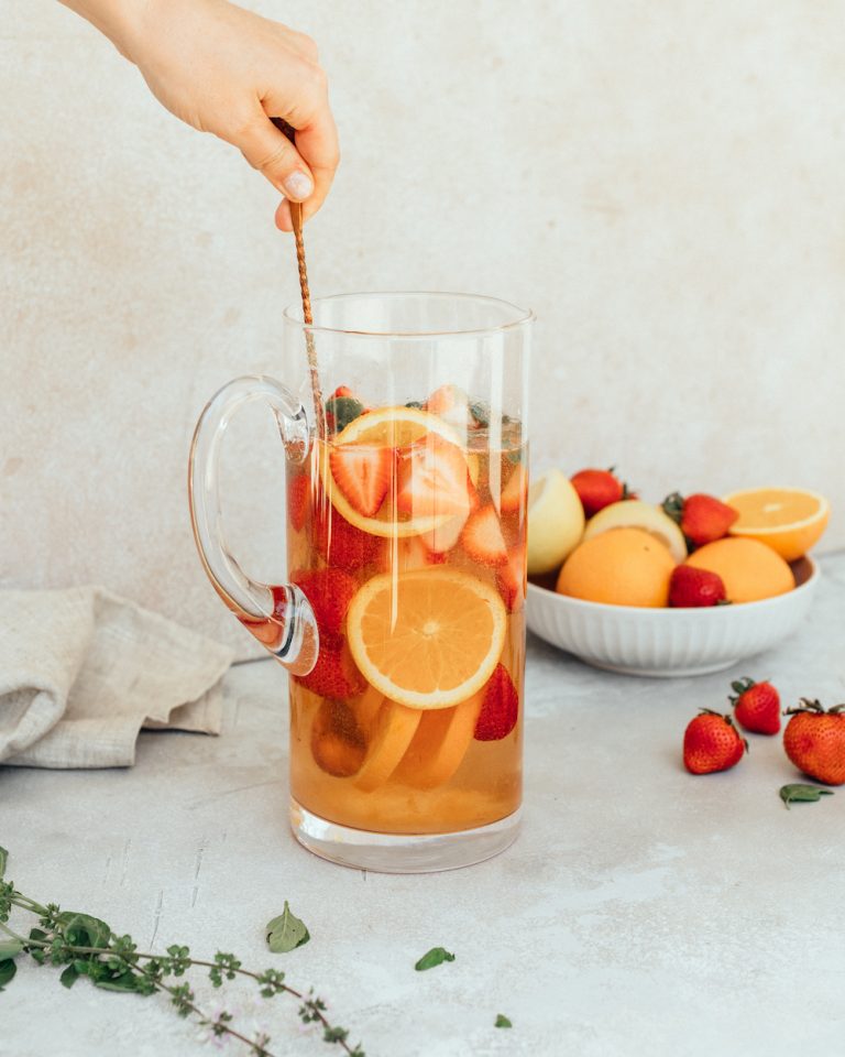 Pitcher of rosé sangria recipe being mixed with a long copper spoon next to a bowl of fruit with strawberries and oranges.