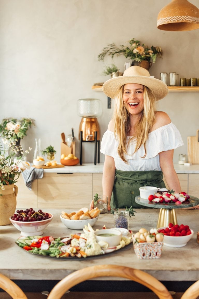 Blonde woman wearing hat smiling behind table of appetizers.