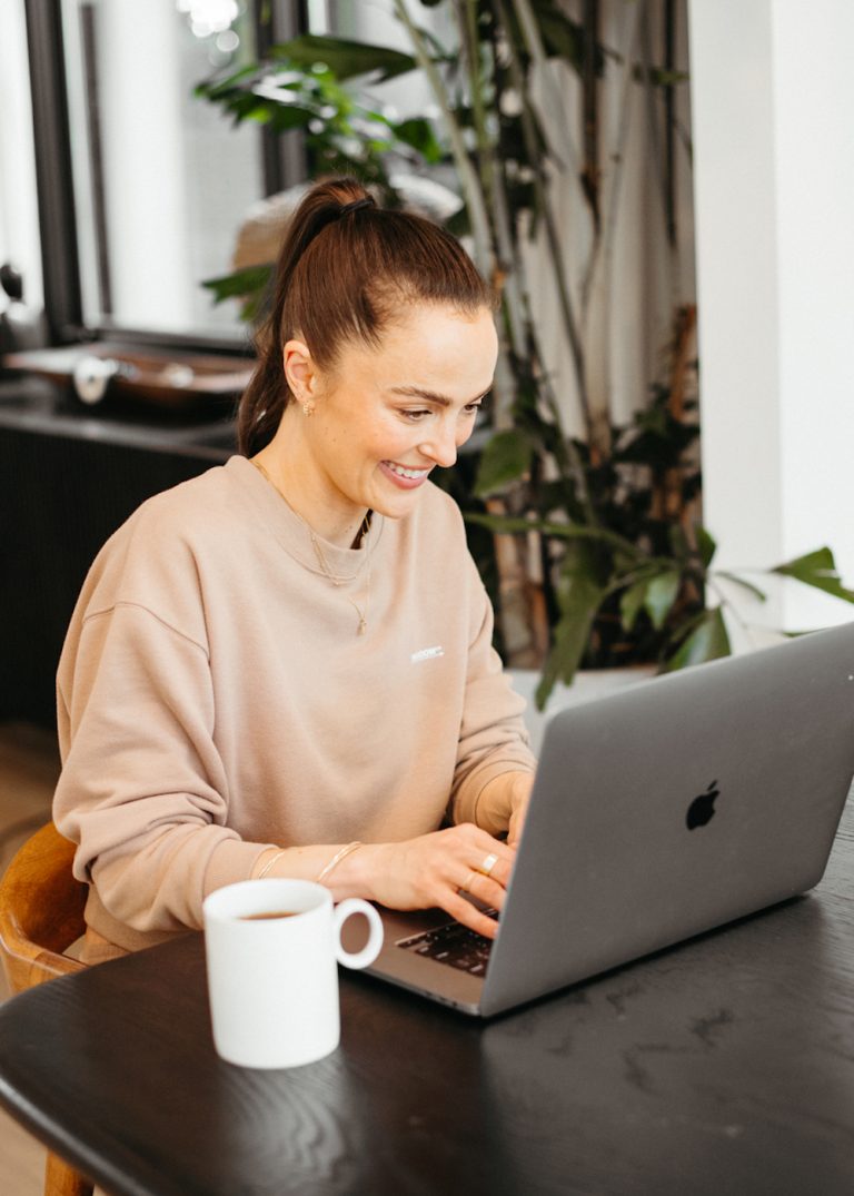 Brunette woman typing on Apple laptop at desk with white mug of coffee.