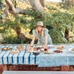 Blonde woman wearing sun hat setting out food dishes on outdoor dining table.