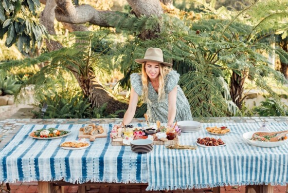 Blonde woman wearing sun hat setting out food dishes on outdoor dining table.