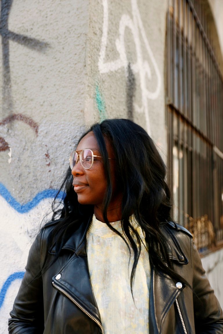 Black woman wearing leather jacket and light yellow dress standing in front of graffiti wall.
