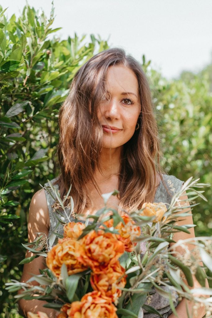 Brunette woman standing in front of trees carrying bouquet of flowers.