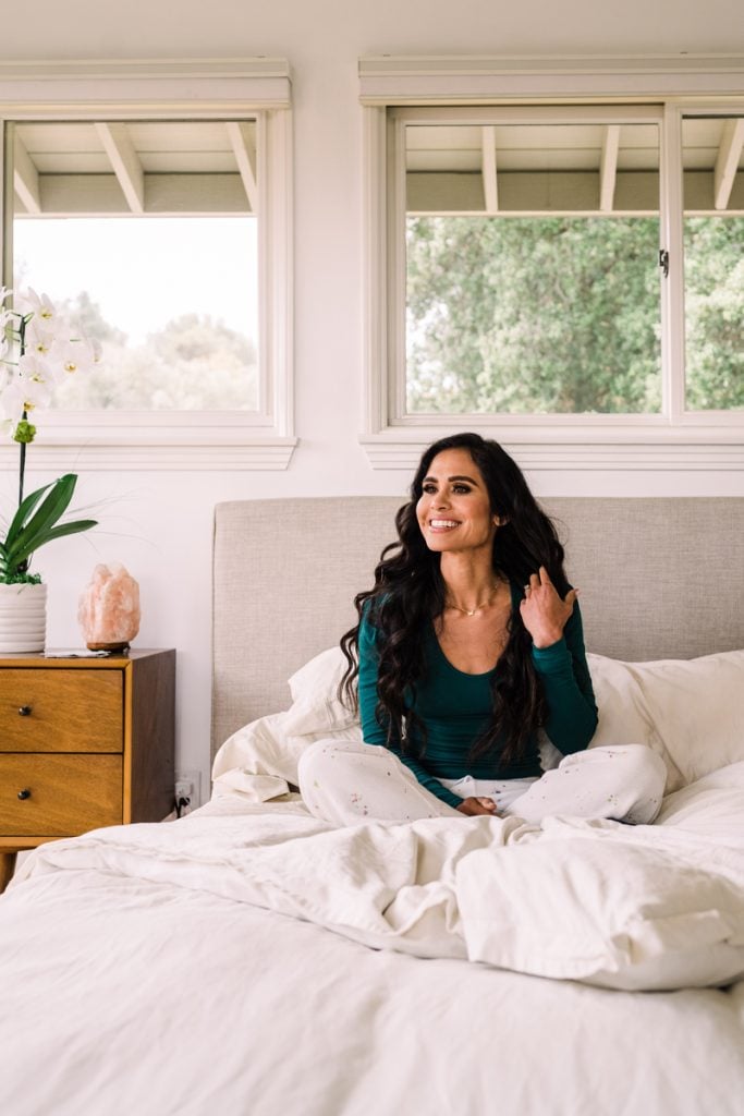 Woman with dark brown hair wearing long-sleeved teal t-shirt lying in bed with white sheets.