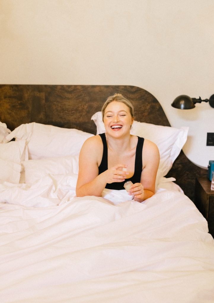Blonde woman holding supplement bottle lying in bed wearing black tank top.