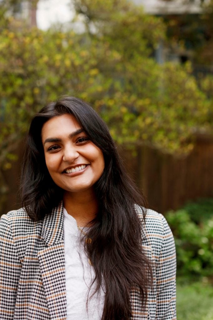 Indian woman with long brown hair smiling wearing plaid blazer.