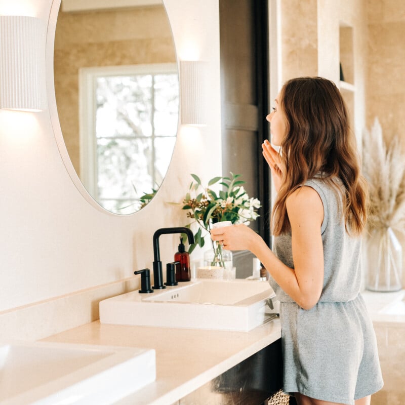 Brunette woman applying skincare lotion in bathroom mirror.