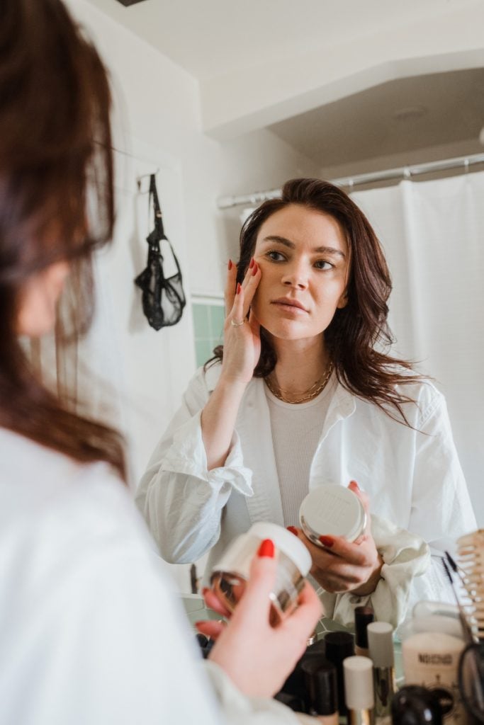 Brunette woman wearing white shirt applying moisturizer in mirror.