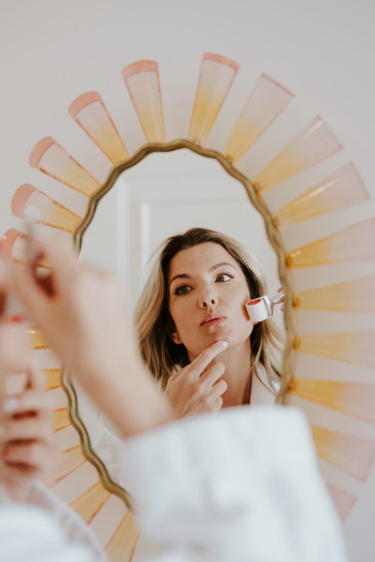 Blonde woman using facial sculpting tool in mirror.