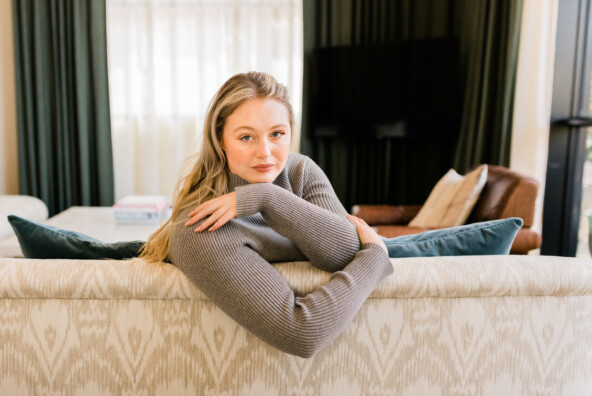 Blonde woman wearing gray sweater leaning over couch.