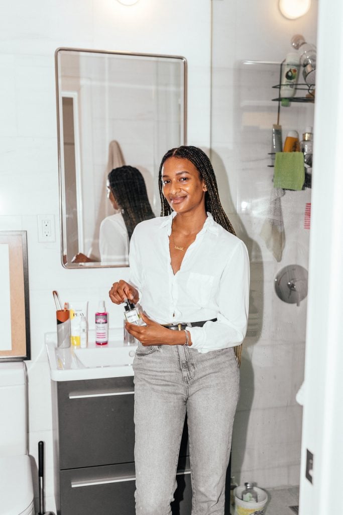 Black woman wearing button-down shirt and jeans using serum in bathroom.