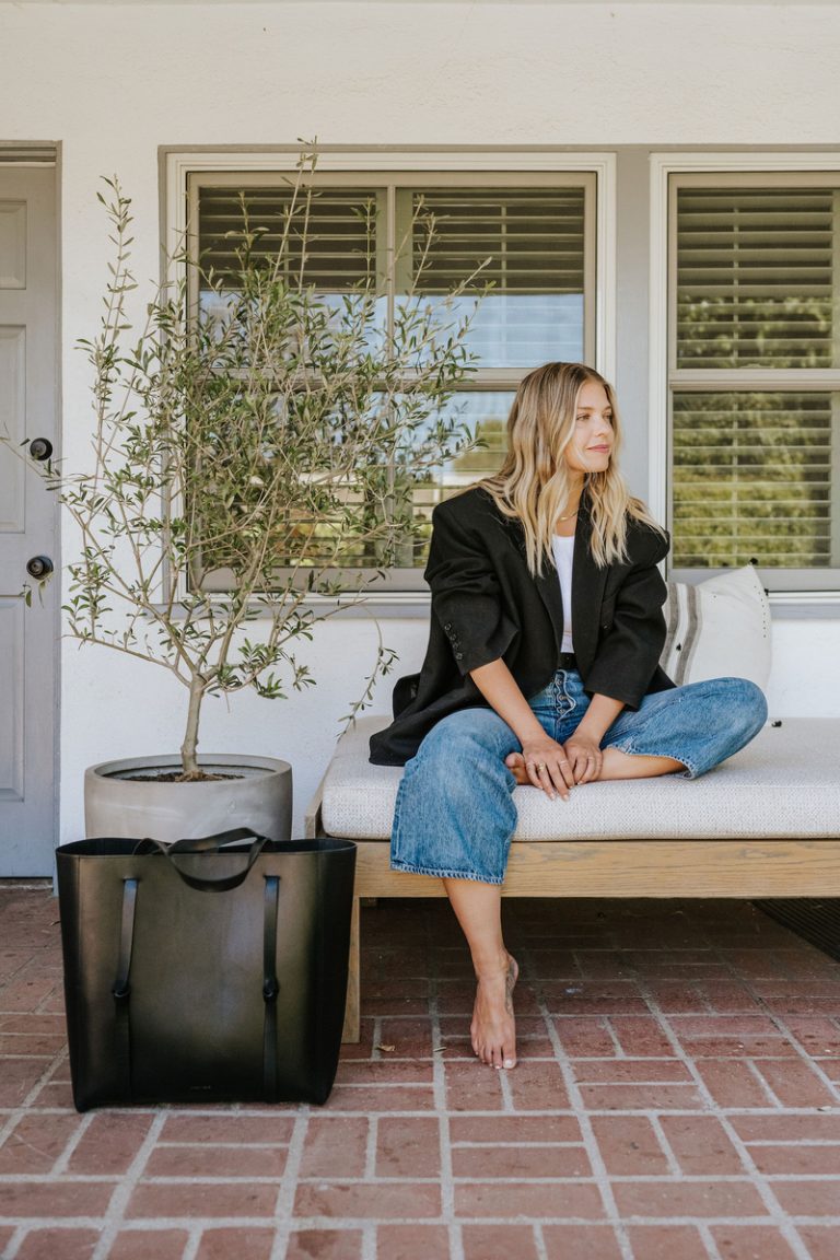 Blonde woman in jeans and black jacket sitting outside.