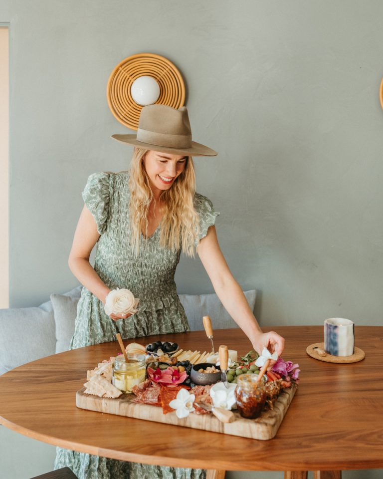 Blonde woman wearing hat arranging ingredients on charcuterie board.