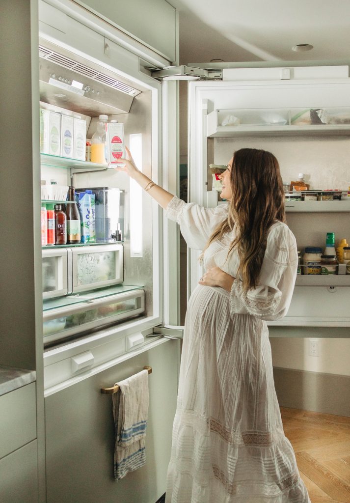 Pregnant woman wearing long white dress opening fridge in kitchen.