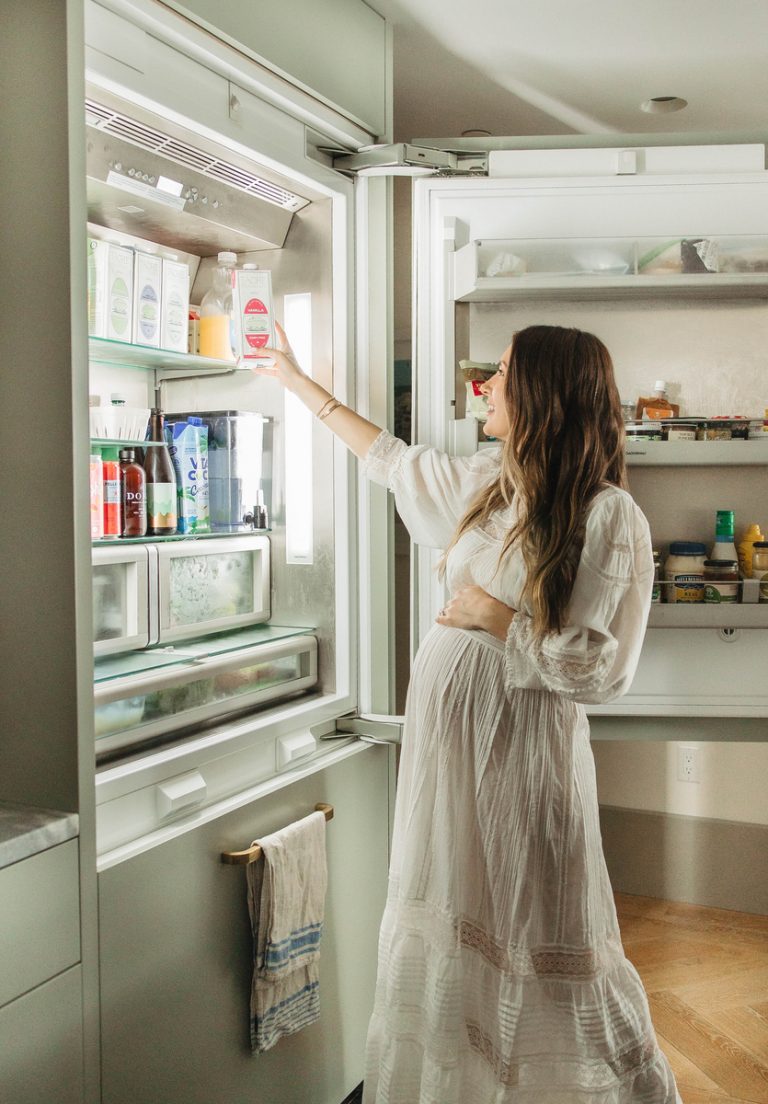 Pregnant woman wears long white dress and opens fridge in kitchen.