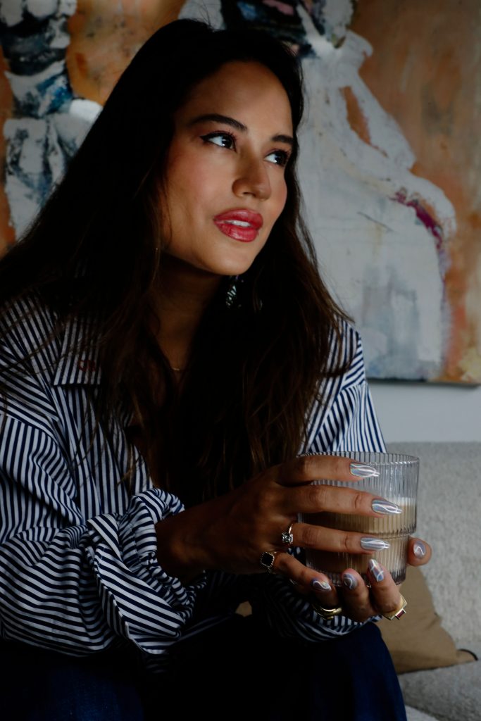 Brunette woman wearing striped blue and white shirt holding glass cup of coffee.
