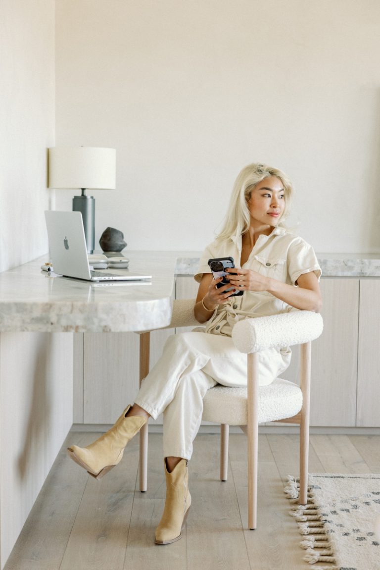 Blonde Asian woman wearing white jumpsuit and cowboy boots holding phone sitting at desk.