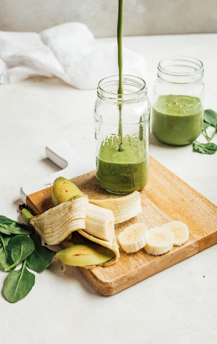 Pouring green smoothie into mason jar on cutting board with sliced up banana.