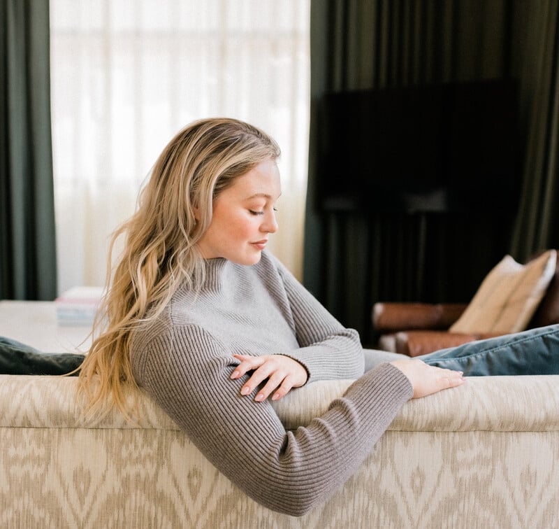 Blonde woman leaning over couch wearing gray sweater.