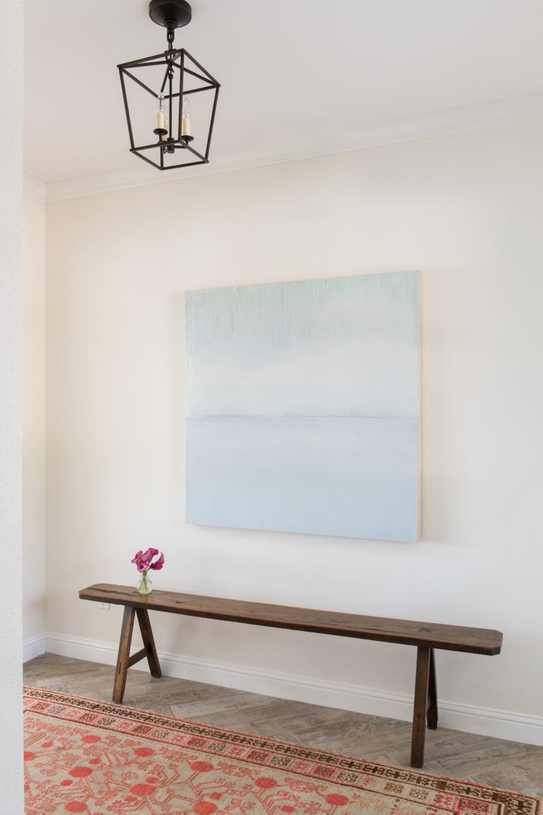 White minimalist foyer with white and aqua abstract painting, wooden bench and oriental rug.