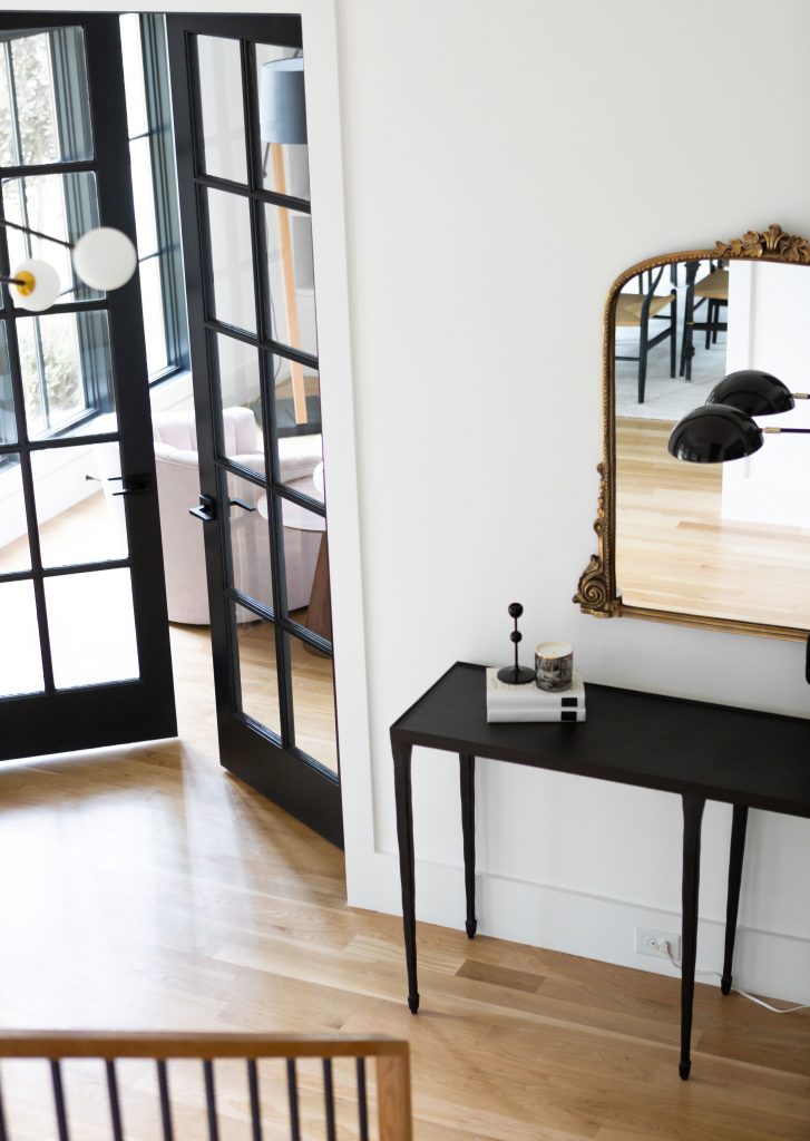 White entryway with wood floors, black French doors, black console table, and gilded mirror.