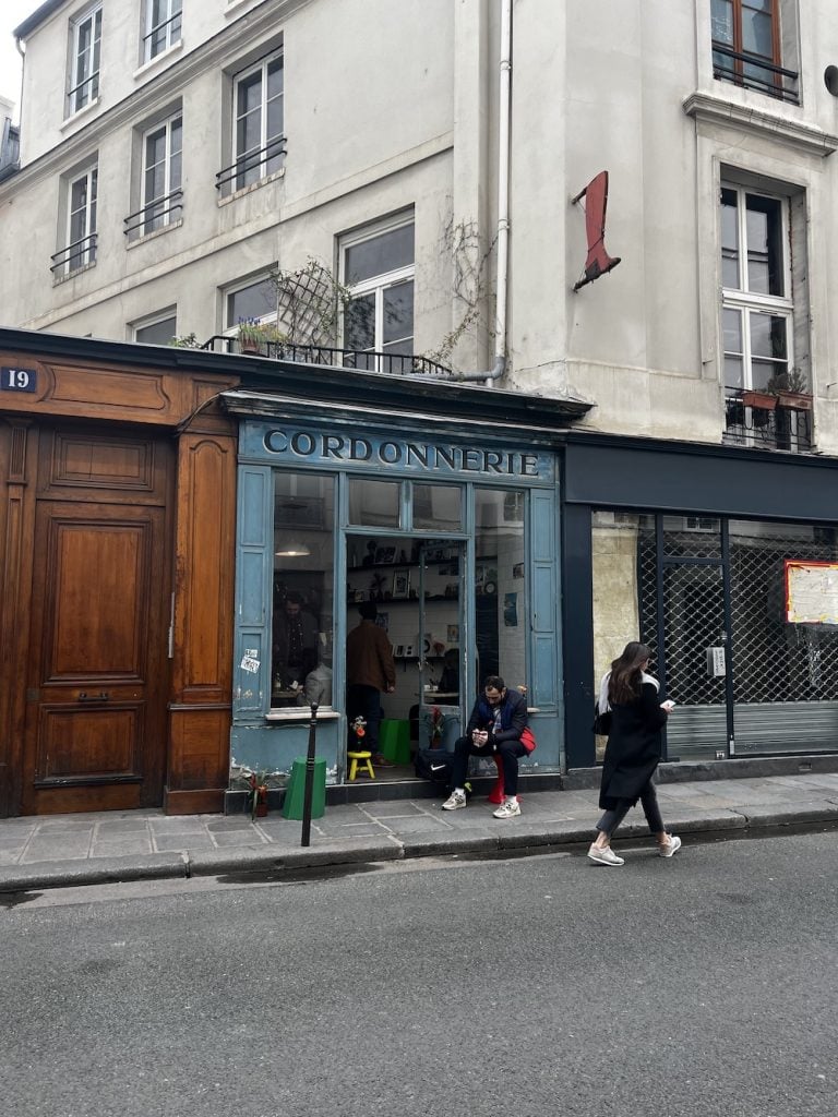 A man sitting outside of Boot Café while a woman walks by in Paris.