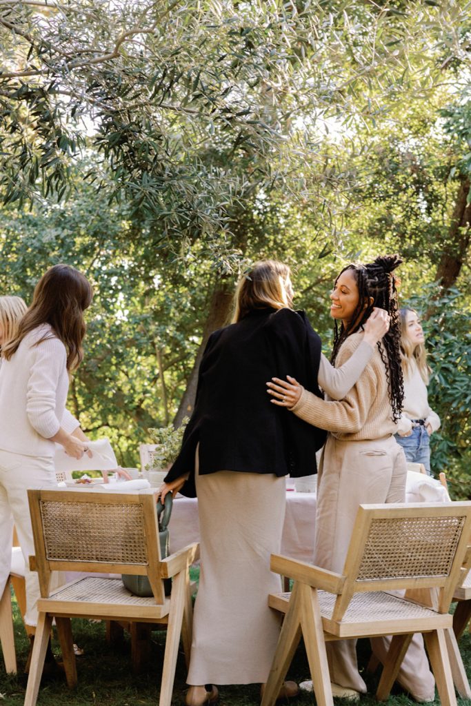Two women hugging at outside dinner table surrounded by three other women. Friendships after kids.