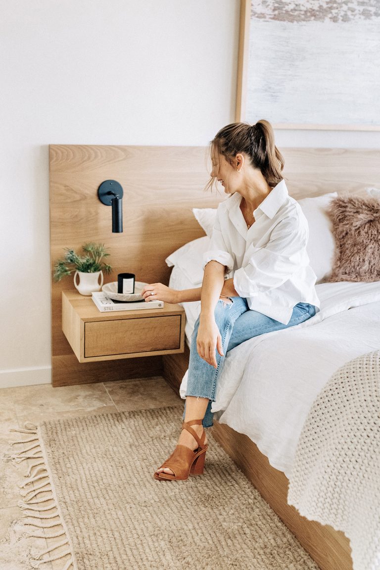 Brunette woman with white sheets sitting on bed with her arms crossed.