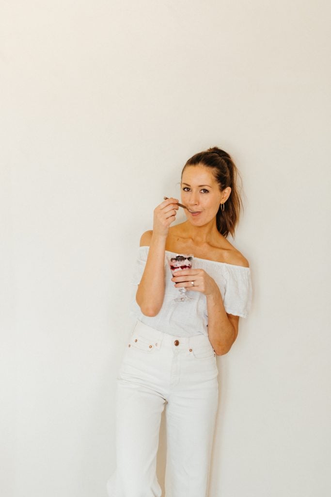 A brunette woman wearing a white shirt and jeans eating a parfait dessert.