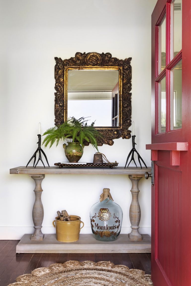 White entryway with red front door, wooden console and gilded mirror.