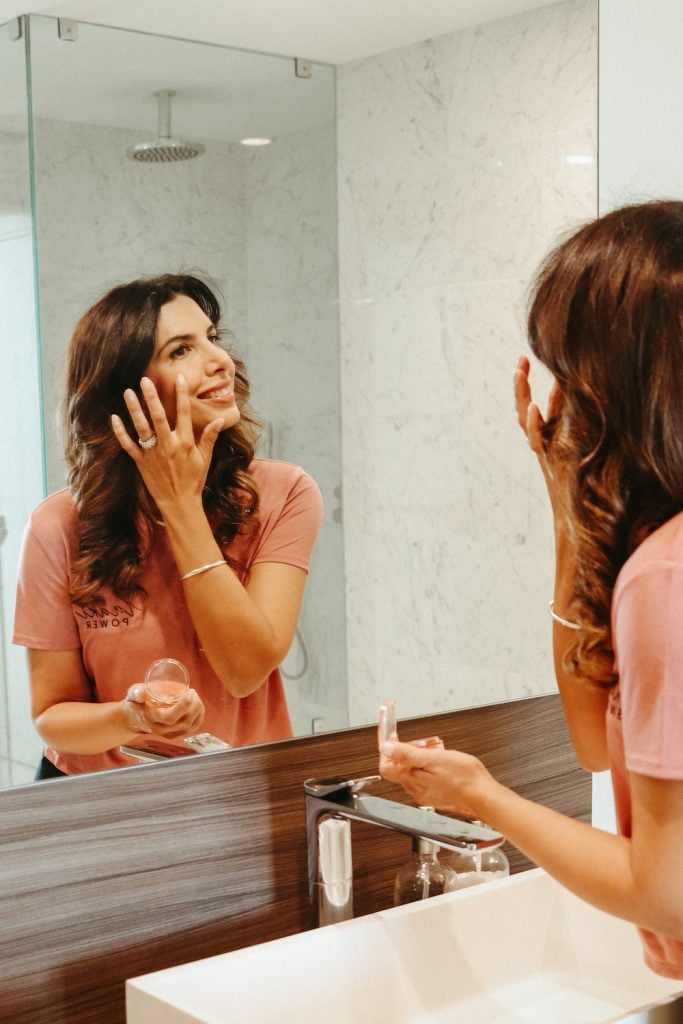 Brunette woman in pink shirt applying makeup in bathroom mirror.