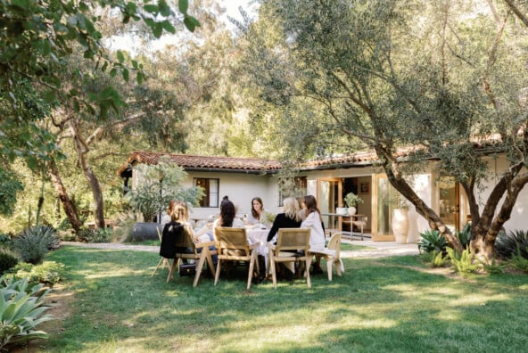 Women sitting around outdoor dining table in the middle of green grass yard.