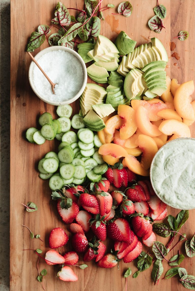 Salad greens, avocado, peach, cucumber, strawberry, salt and sauce arranged on a wooden cutting board.