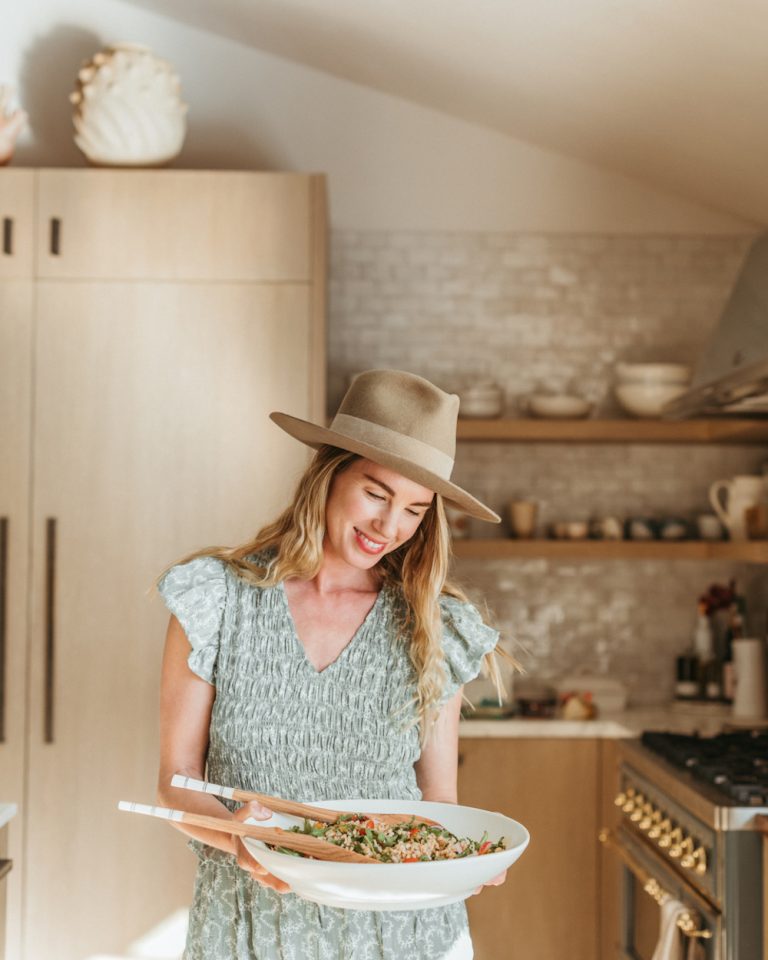 Blonde woman wearing blue dress and sun hat holding white bowl of salad in the kitchen.