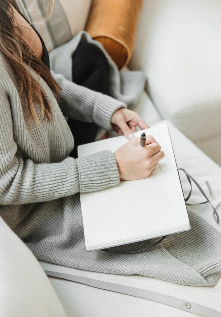 Woman wearing long gray sweater journaling on couch.