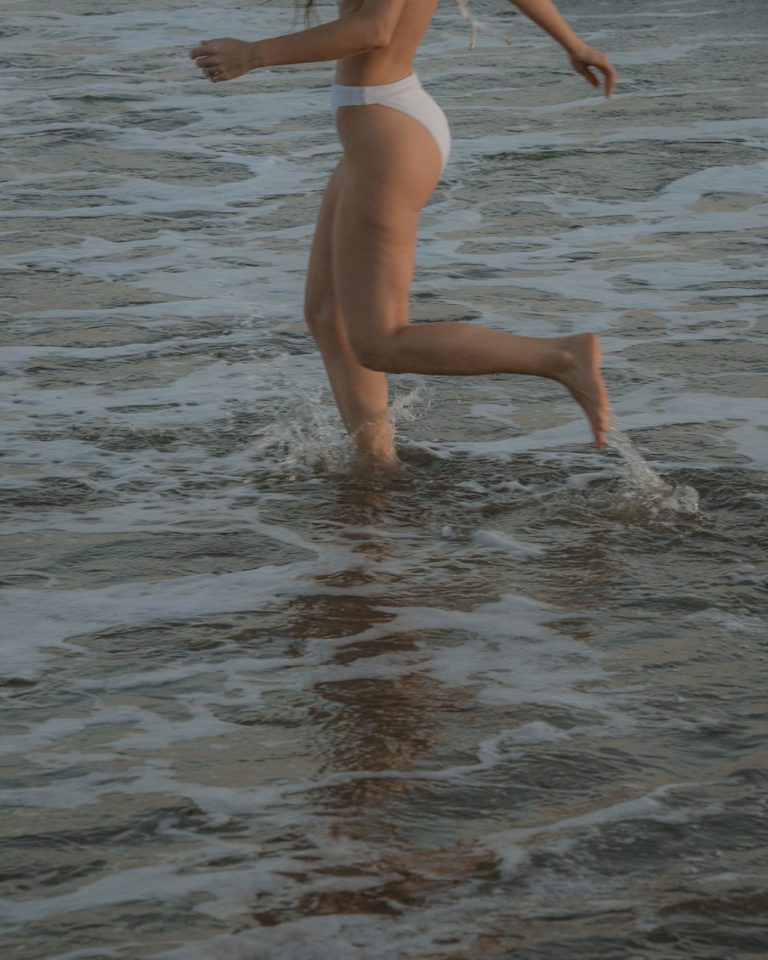 Woman jumping in ocean wearing bathing suit.