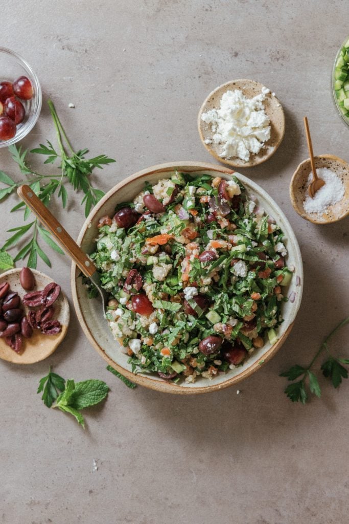 Colorful grain salad surrounded by pinch bowls of olives, feta cheese, and salt.