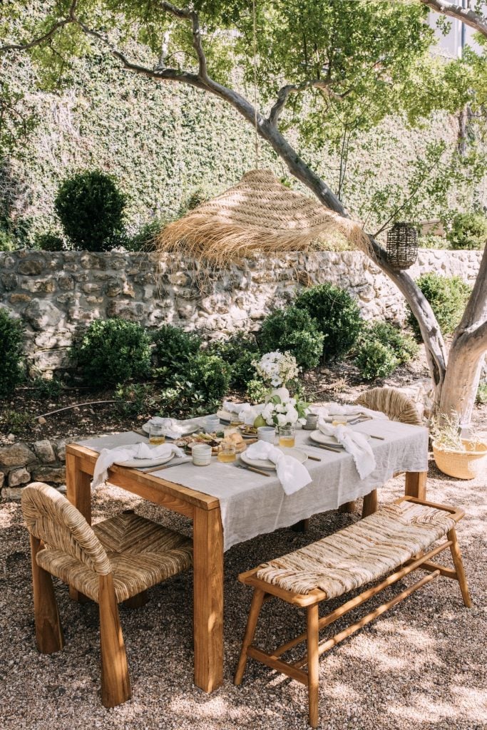Outdoor dining table with two chairs and bench set with linen tablecloth and neutral table settings.