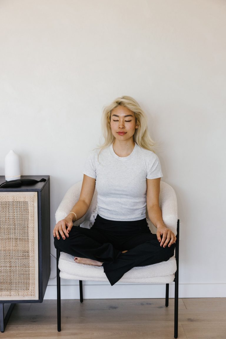 Blonde woman wearing gray shirt and black pants meditating in chair.