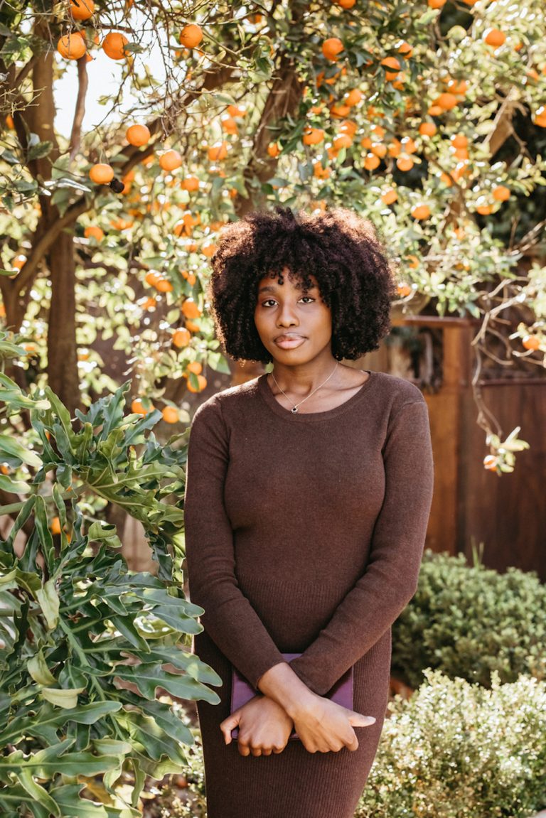 Black woman in a brown dress standing by an orange tree and holding a diary.
