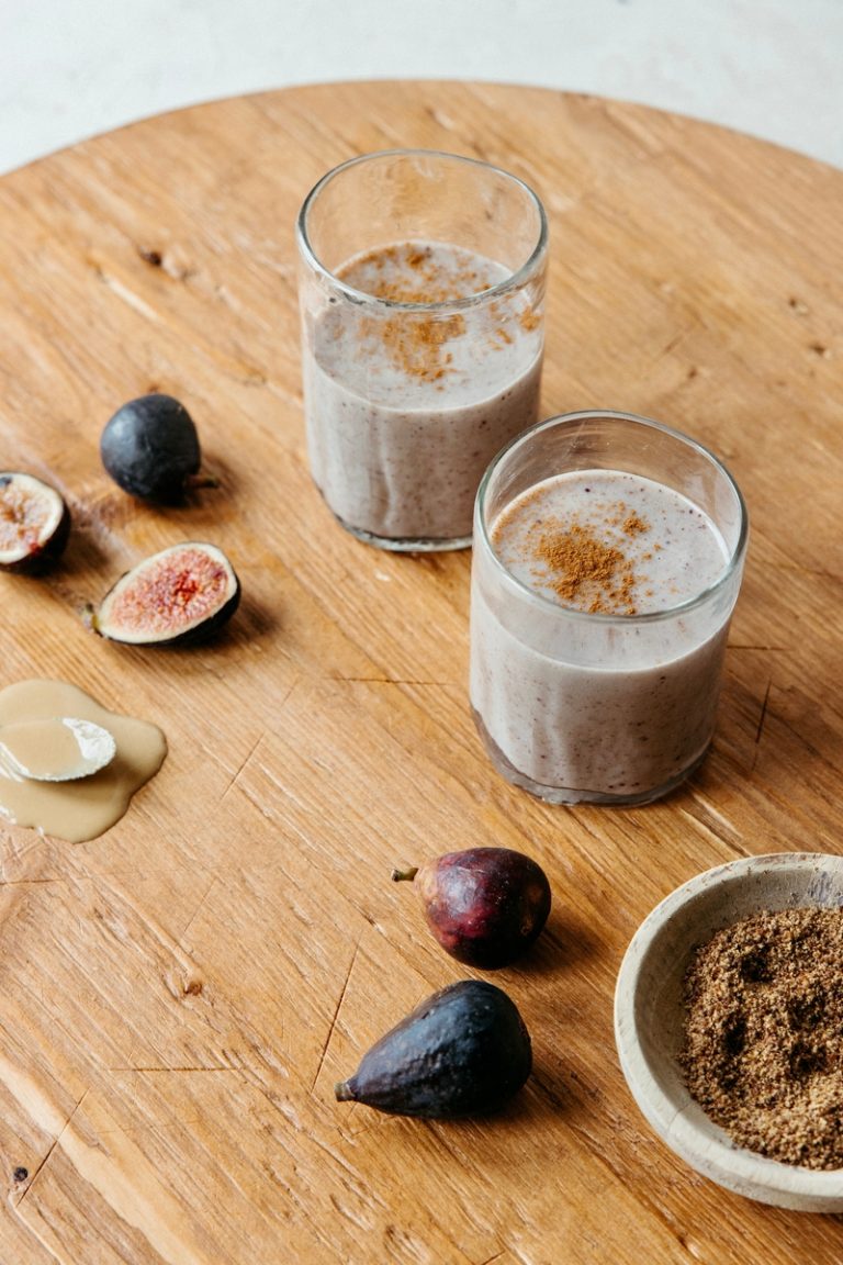 Two glasses of fig smoothie sprinkled with cinnamon surrounded by sliced figs on wooden board.