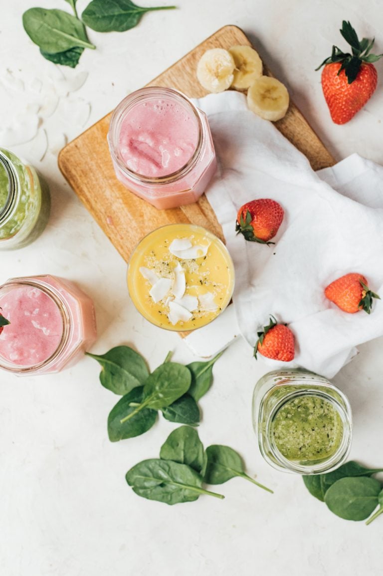 Pink, green, and yellow smoothies on a marble countertop with spinach, strawberries, and bananas.