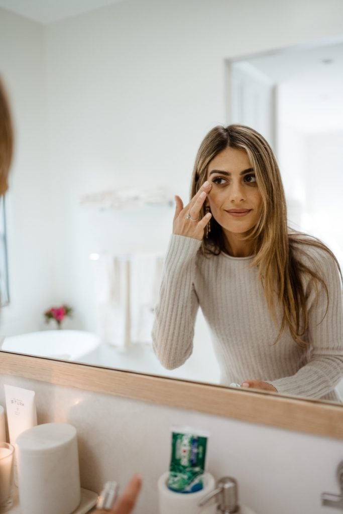 Woman applying makeup under eyes in mirror.