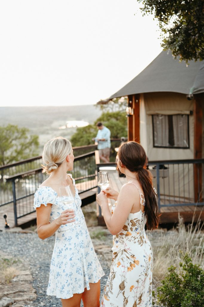 Two women holding white wine looking behind them at yurt and field.