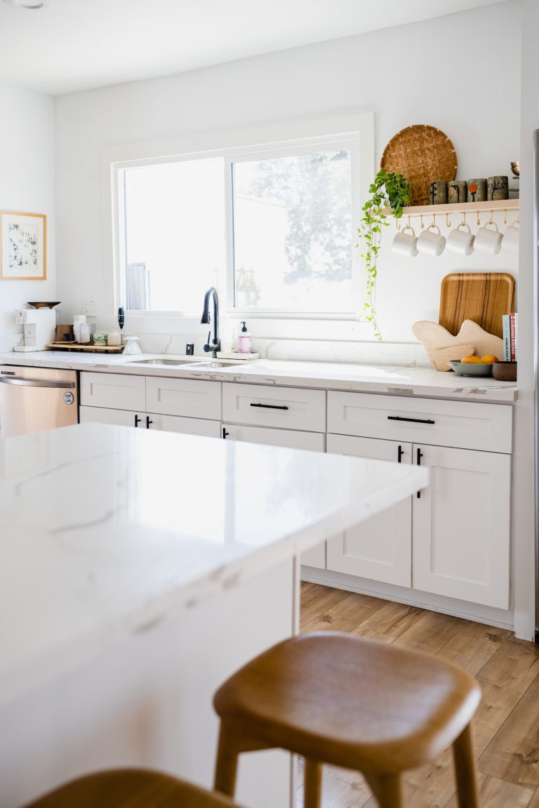 A bright, white kitchen with marble countertops, a window behind the sink, and natural shelving with draped plants and white mugs.